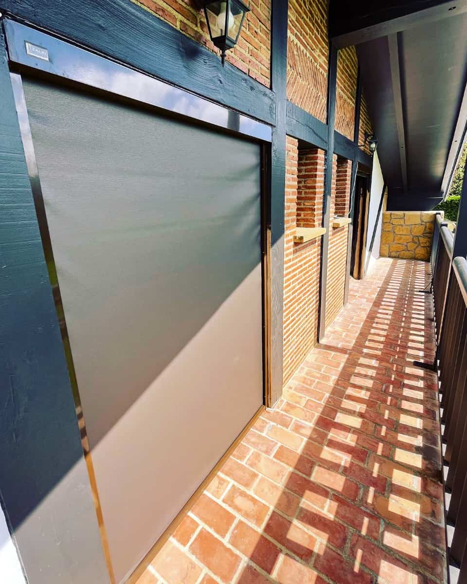 Screen motako toldo bertikala, adreilu gorrizko horma eta egur habeak dituen estilo rustikoko balkoi batean instalatua, Itzalki Toldoak enpresak Zarautzen (Gipuzkoa, Euskal Herria), Aian ere zerbitzua eskaintzen duena