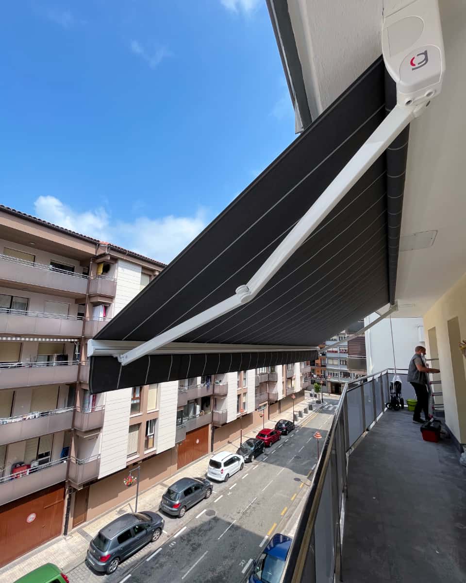 Marra beltz eta zuriak dituen toldo hedagarria, hiri-inguruneko etxebizitza bateko balkoian instalatua, Itzalki Toldoak enpresak Zarautzen (Gipuzkoa, Euskal Herria) egindakoa, Usurbilen ere zerbitzua eskaintzen duena